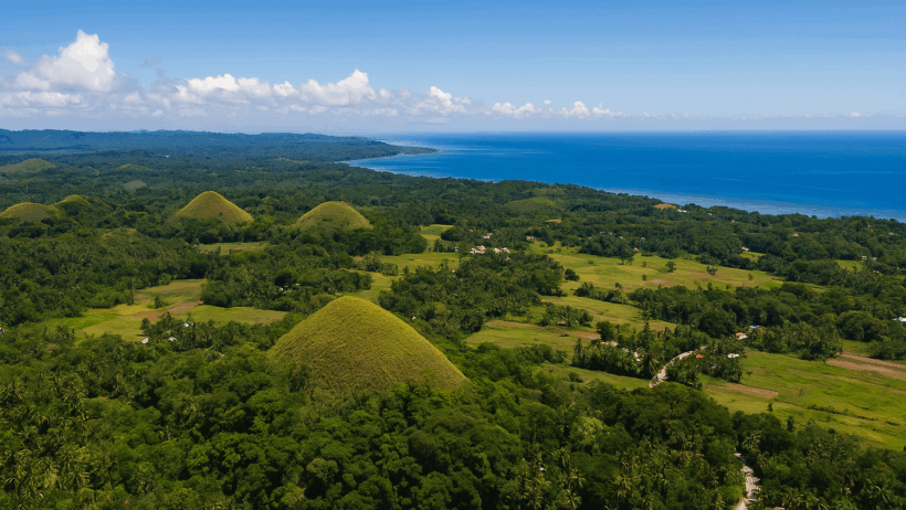 Aerial view of Bohol’s famous Chocolate Hills surrounded by lush forests, farmland, and a distant blue coastline under a clear sky.