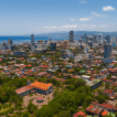 Aerial view of Cebu City showing its modern skyline, historic landmarks, and coastal waters, with green parks and urban neighborhoods in the foreground under a bright blue sky.