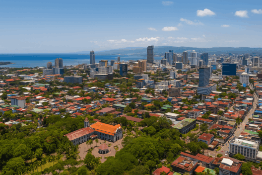 Aerial view of Cebu City showing its modern skyline, historic landmarks, and coastal waters, with green parks and urban neighborhoods in the foreground under a bright blue sky.
